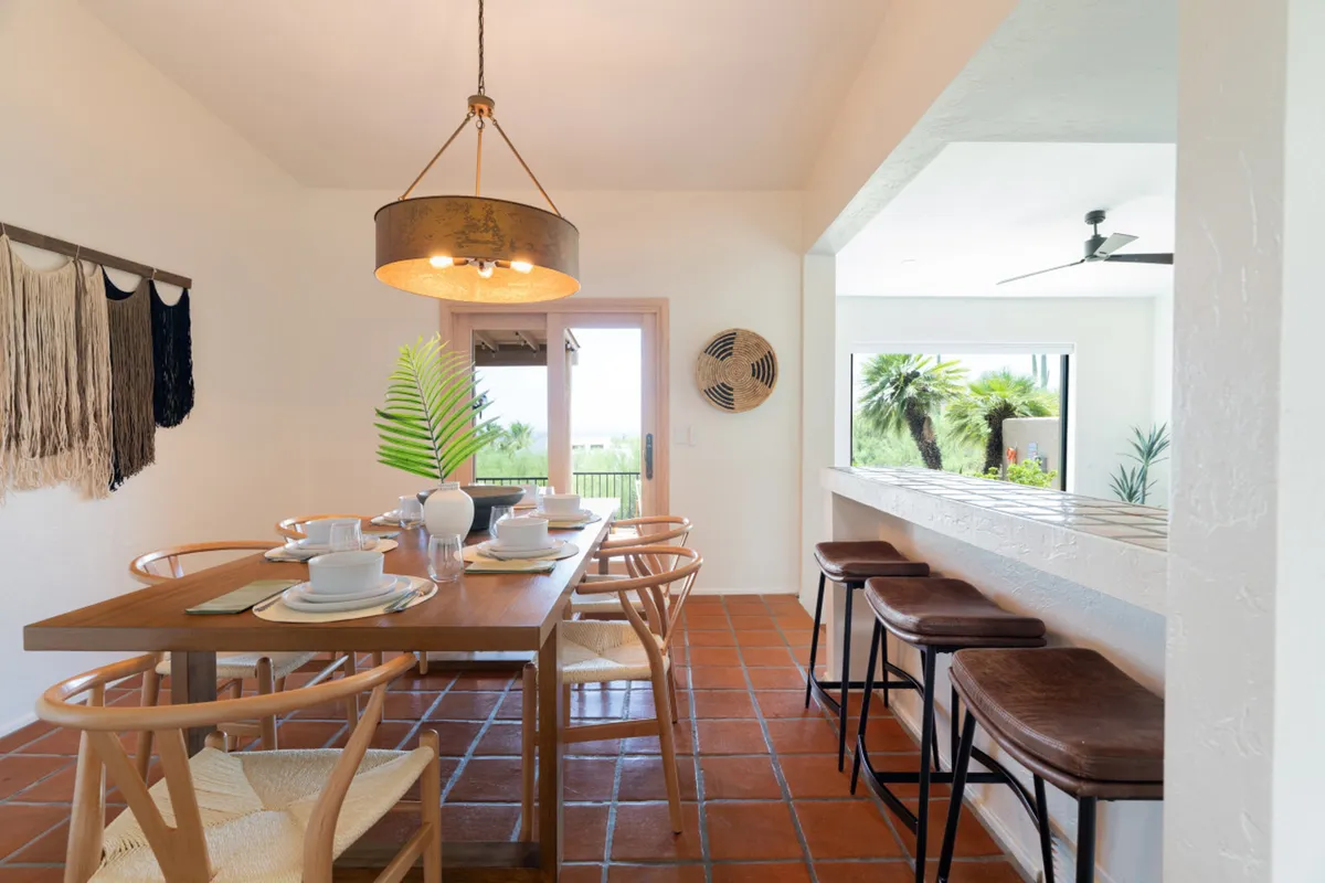 A dining area featuring a table set with dishes, a stylish hanging light fixture, and decorative greenery. Large windows allow for natural light and glimpses of the outdoors, creating a warm, sociable environment ideal for shared meals and gatherings.