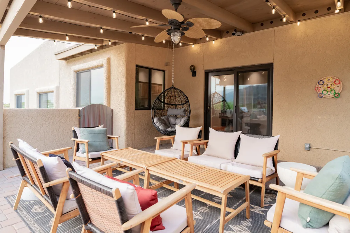 A charming exterior lounge area with a hanging chair, a wooden coffee table, comfortable seating, and decorative wall art illuminated by string lights. The enclosed, thoughtfully styled space feels both secure and intimate, perfect for evening relaxation or casual gatherings. The design balances openness to nature with a sense of privacy.