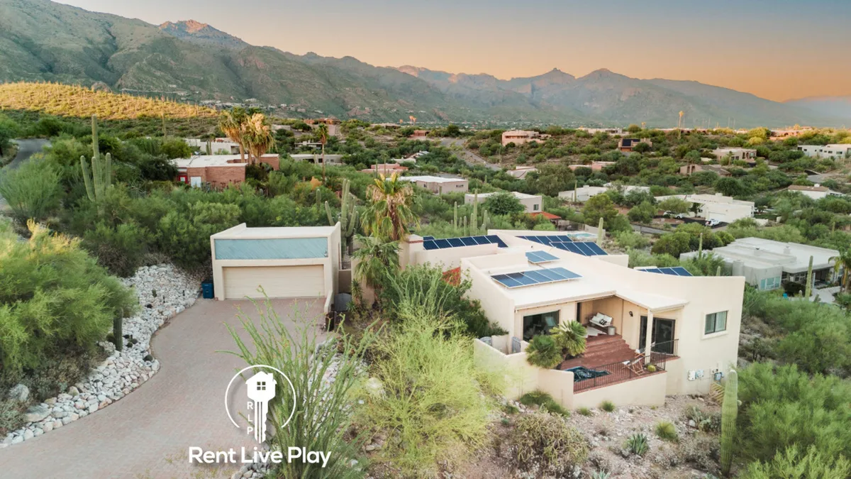 This exterior image shows the house situated amidst a stunning desert landscape, with solar panels on the roof and mountains in the background. The natural terrain and cacti convey the quintessential Sonoran Desert environment, inviting guests to connect with the outdoors and explore the surrounding trails.