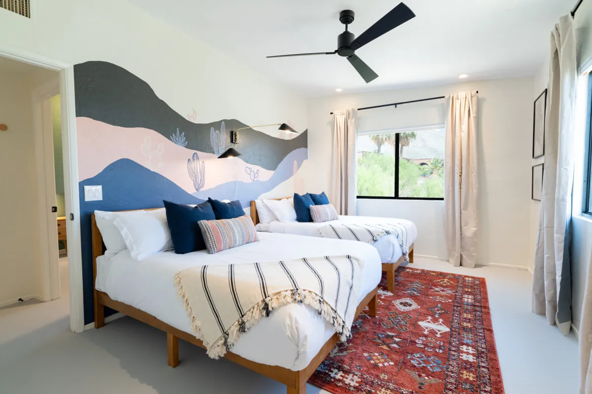 A welcoming guest bedroom with two beds dressed in white linens and accented with colorful pillows, a decorative wall mural, an area rug, and a large window allowing for ample sunlight. The room feels cozy and flexible, ideal for families or friends sharing the space.