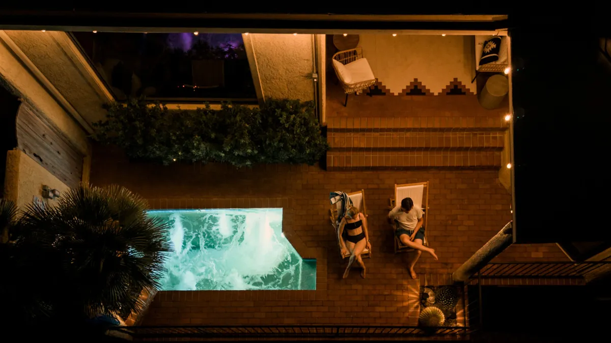 A tranquil swimming pool scene with two lounge chairs set beside the water, surrounded by lush greenery and illuminated by the soft glow of string lights as evening falls. The peaceful atmosphere invites relaxation and socializing under the stars.