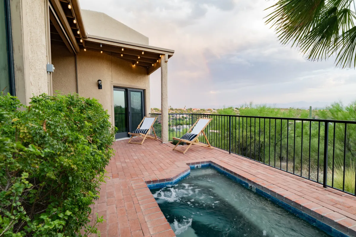 An inviting view of the custom in-ground heated 'spool' (hot tub/pool combo) surrounded by lounge chairs and lush desert landscaping. The area is bathed in natural light with open skies above, suggesting opportunities for both daytime relaxation and evening soaks under the stars. The space is clearly designed for comfort and socializing, with ample seating and a tranquil vibe.