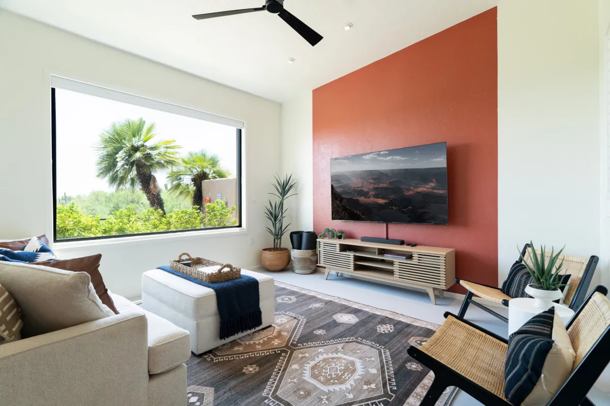 A cozy and inviting living room with a comfortable seating arrangement centered around a modern TV on a wooden stand. A large window floods the room with natural light and offers a lush, green view, while a geometric patterned rug and decorative details enhance the welcoming ambiance.
