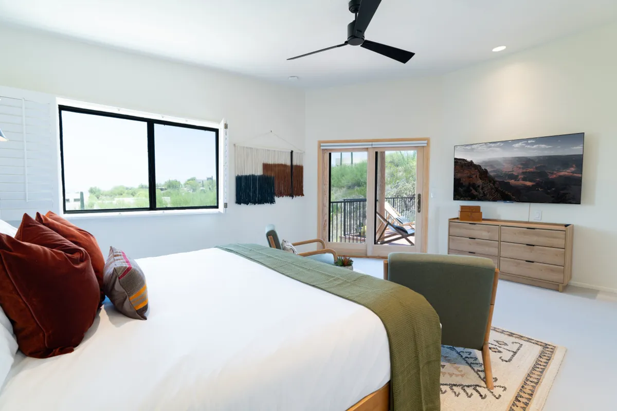 A serene bedroom featuring a bed with decorative pillows and a plush green throw blanket, large windows that flood the space with natural light, a wooden dresser, and a wall-mounted television. The room exudes comfort and relaxation, perfect for unwinding after a long day.