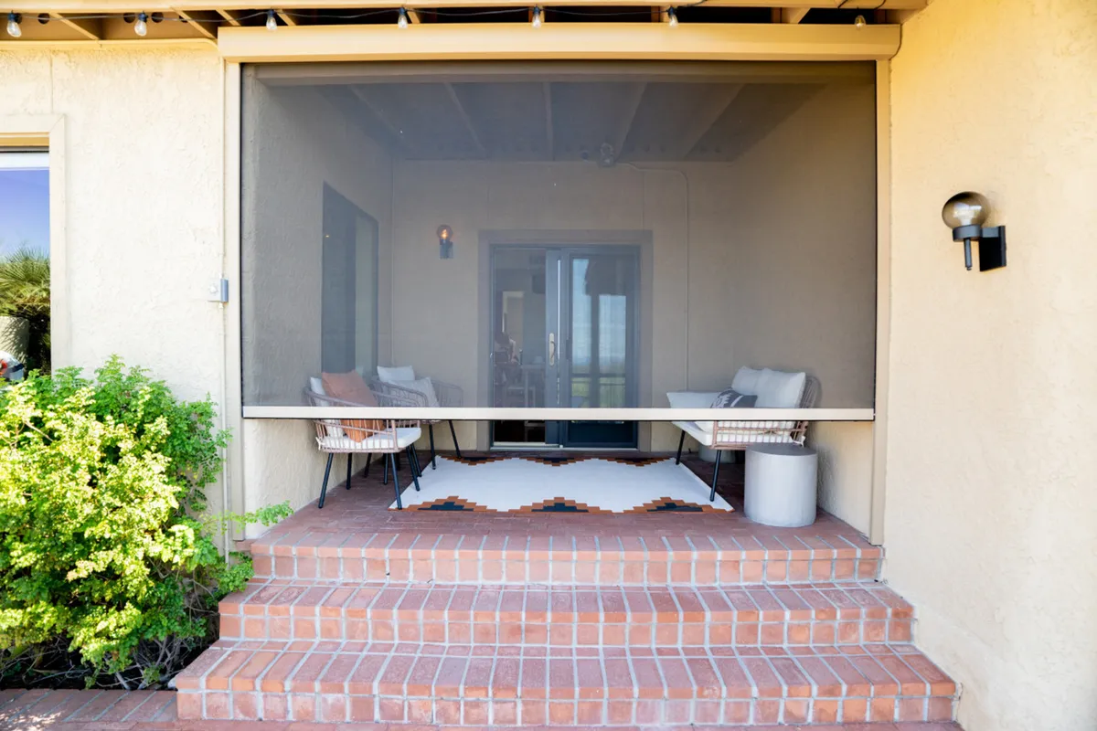This image features steps leading up to the entrance of the property, flanked by two outdoor chairs and a decorative rug. The porch area is inviting and open, set against a backdrop that suggests a blend of desert elements and serene outdoor living. The scene evokes the atmosphere of Arizona's desert landscape, with a focus on relaxation and observing the natural surroundings from a comfortable vantage point.