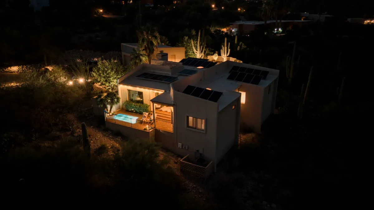 An illuminated patio area featuring a swimming pool, surrounded by cacti and desert landscaping. Solar panels on the roof underline the home's modern amenities, while the cozy lighting sets the mood for intimate outdoor gatherings.