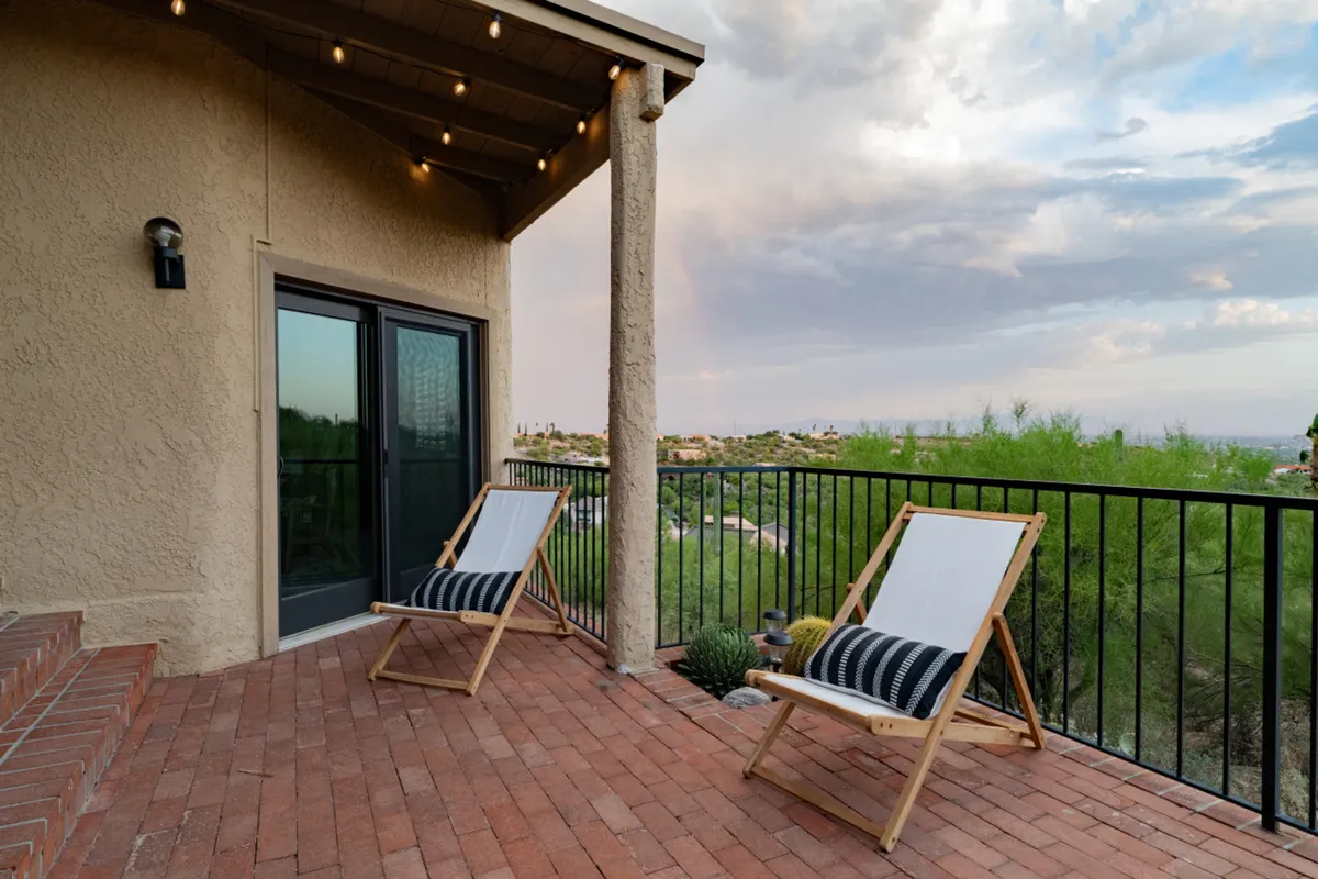 A pair of wooden lounge chairs are positioned on a terrace overlooking a sweeping natural landscape. The setting sun casts a golden glow, creating a serene backdrop for relaxation and contemplation.