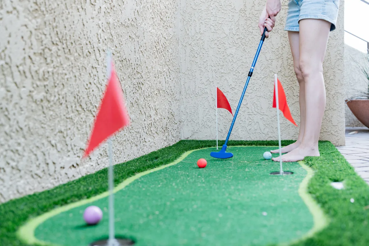 An exterior scene showcasing a putting green framed by desert landscaping, with a golf club, vibrant balls, and red flags visible. The background includes natural greenery, emphasizing outdoor activity and enjoyment of the spacious grounds. The playful setup hints at family fun and the chance to explore the property’s acre of land.